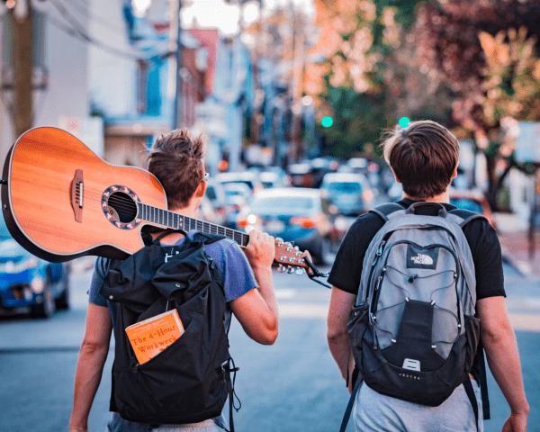 Two backpackers walking on the road side by side.The person on the left is holding a guitar over their shoulder.